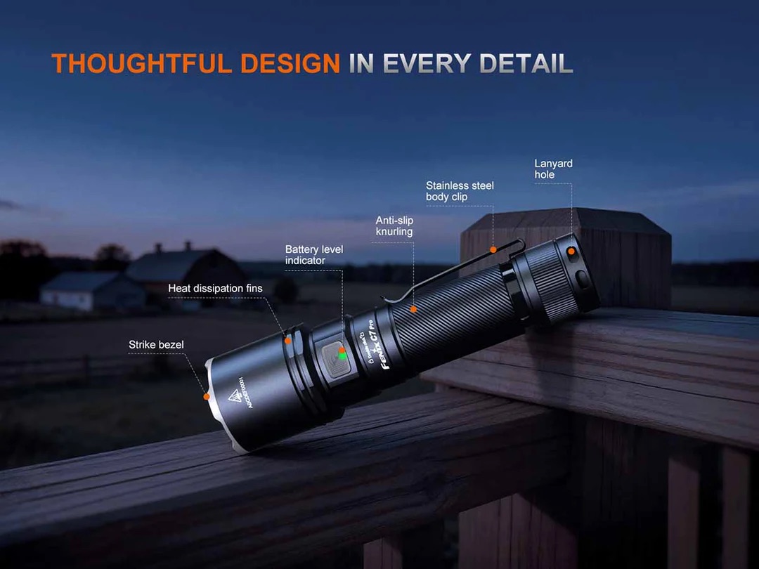 Close-up of a tactical flashlight resting on a wooden railing with labeled parts: strike bezel, heat-dissipation fins, battery level indicator, anti-slip knurling, stainless steel body clip and lanyard hole.